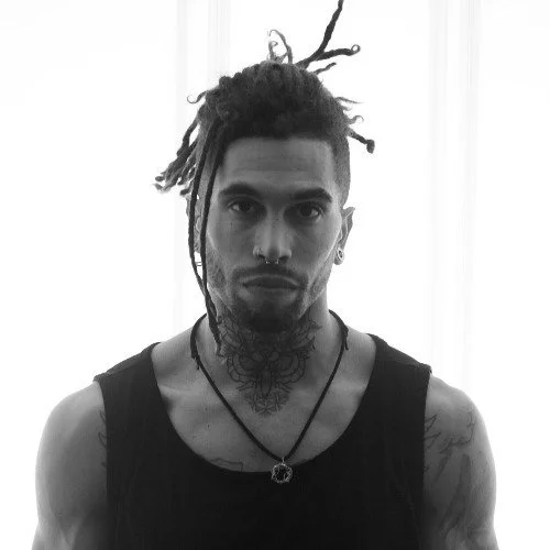 Black and white photo of a man with dreadlocks, tattoos, and facial piercings, wearing a sleeveless shirt and necklaces, standing in front of a light background.