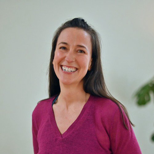 a woman with long brwon hair and a pink vneck jumper smiling with a grey wall and plant in the background