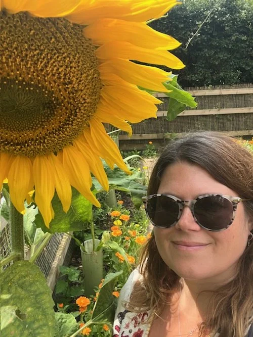 A woman with brown hair wearing sunglasses smiling in a garden with a large sunflower in the foreground and various flowers and greenery in the background.