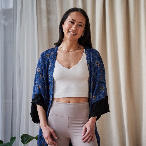 Young woman standing in front of beige curtains, smiling, wearing a white crop top, beige pants, and a blue patterned kimono-style robe.