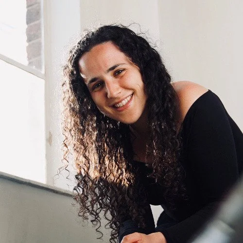 A woman with long curly hair smiling at the camera, wearing a black off-the-shoulder top, sitting indoors near a window.