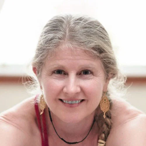 A smiling middle-aged woman with gray hair, wearing large earrings and a red top, sitting indoors with a blurred background.
