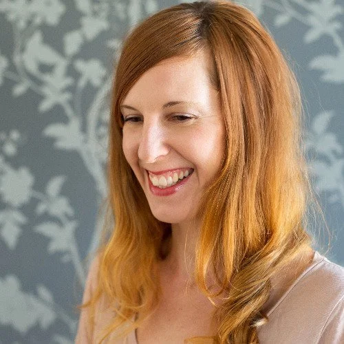 A woman with long red hair and a light beige top, smiling and looking down against a floral patterned wallpaper background.