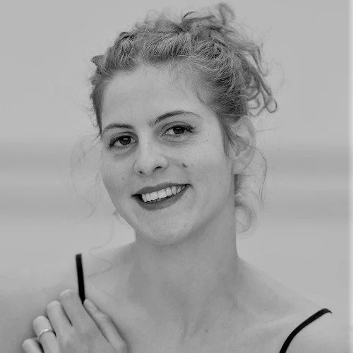 Black and white photo of a woman smiling and holding a cigarette, with her hair tied up in a messy bun, wearing a spaghetti strap top.