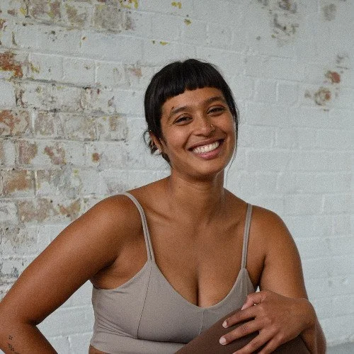 A woman with dark hair tied up and a fringe smiles with a distressed brick wall behind