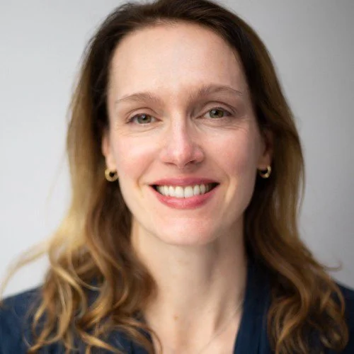A woman with light skin, long wavy hair, and gold earrings smiling at the camera against a neutral background.