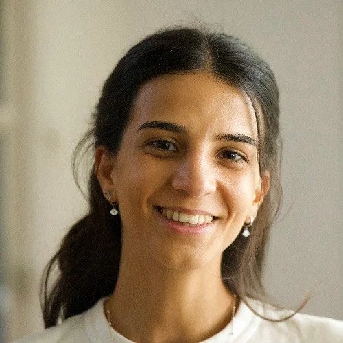 A young woman smiling with dark hair, light skin, wearing earrings and a white top.