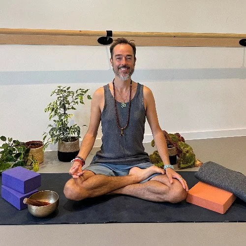 Person practicing meditation in a yoga studio, seated on a yoga mat with their legs crossed and hands resting on their knees, surrounded by plants and yoga props.