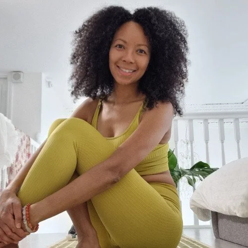 A woman with curly black hair sitting on the floor in a yellow workout outfit, smiling. She is in a bright, cozy room with white walls and a wooden floor, and some greenery is visible in the background.