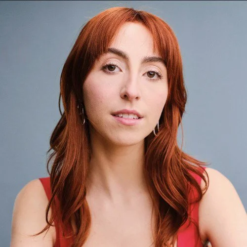 A woman with red hair and a red tank top looks face on with grey background
