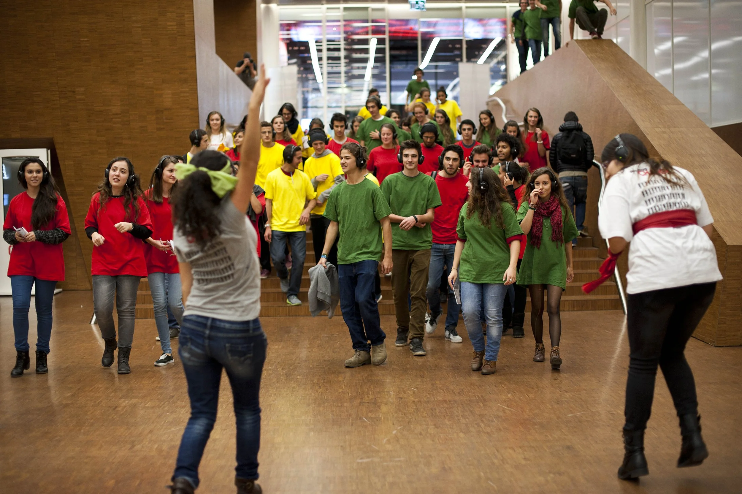 Eine Gruppe von Menschen in roter, grüner und gelber Kleidung geht eine Treppe hinunter, einige tragen Kopfhörer. Die Szene findet in einem modernen Gebäude mit Holzverkleidung statt.