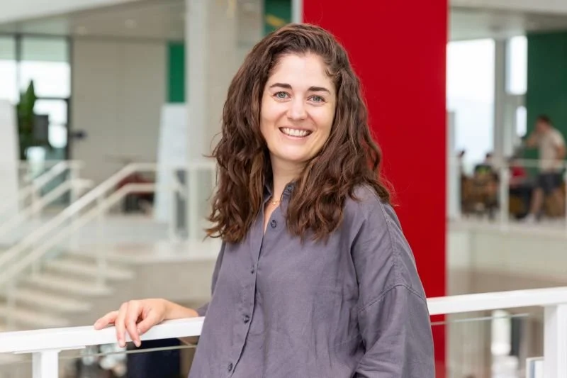 Eine Frau mit lockigem Haar und grauer Bluse lächelt in die Kamera. Im Hintergrund ist ein modernes Büroumfeld mit roten und grünen Elementen sichtbar.