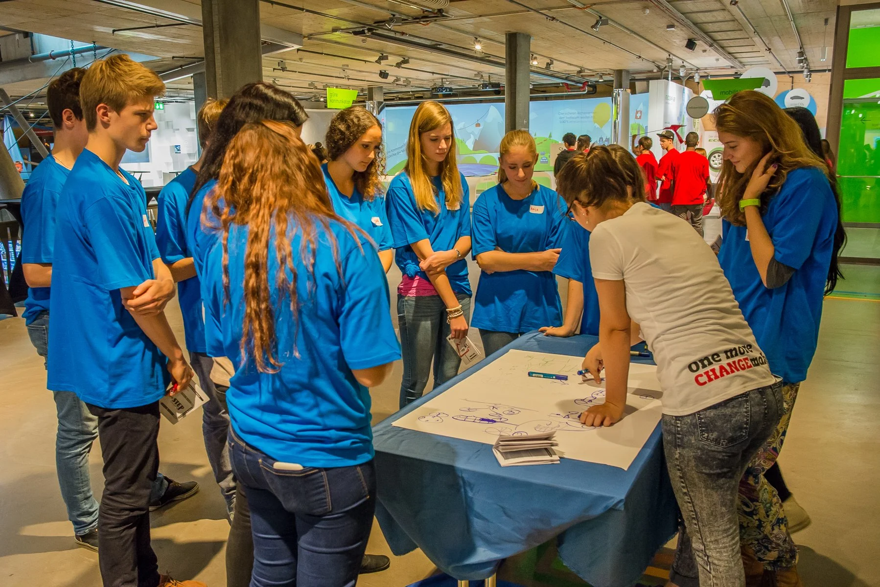 Eine Gruppe von Jugendlichen in blauen T-Shirts steht in einem Raum um einen Tisch, auf dem ein großes Blatt Papier mit Zeichnungen liegt. Eine Person schreibt oder erklärt etwas auf dem Papier, während die anderen aufmerksam zuhören oder zuschauen. Der Raum wirkt modern, mit Betonwänden und heller Beleuchtung.