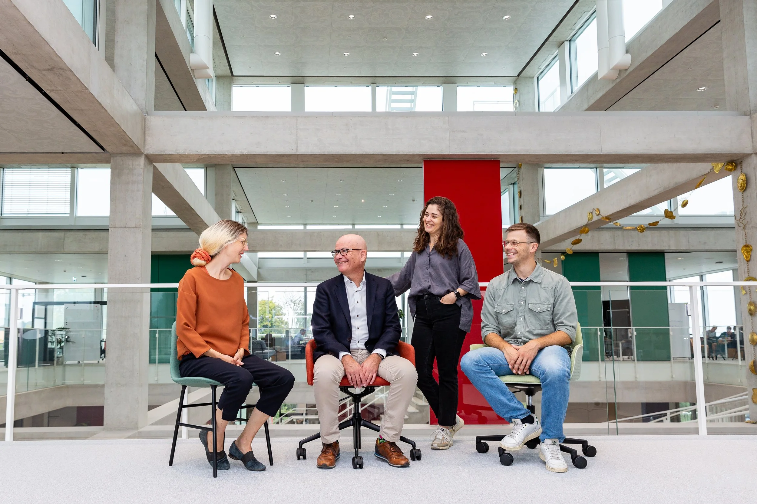 Vier Personen in einem modernen Bürogebäude, lächelnd und auf Stühlen sitzend. Zwei Männer und zwei Frauen. Offene Architektur mit großen Fenstern und einem roten Element im Hintergrund.