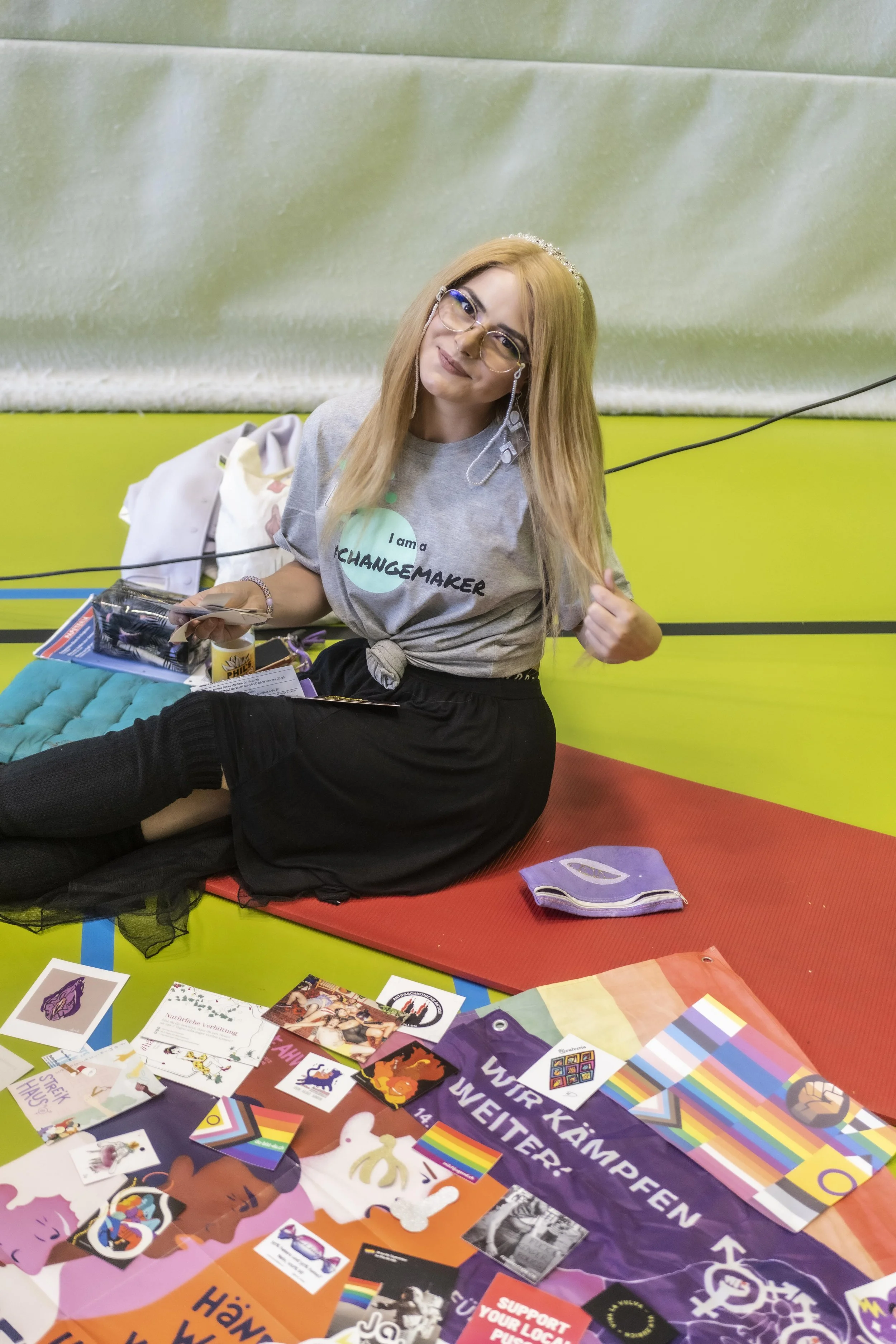 Person mit blonden Haaren und Brille sitzt auf einer Matte, trägt ein graues T-Shirt mit der Aufschrift "I am a changemaker". Umgeben von bunten Flyern und Stickern mit Regenbogenmotiven auf dem Boden.
