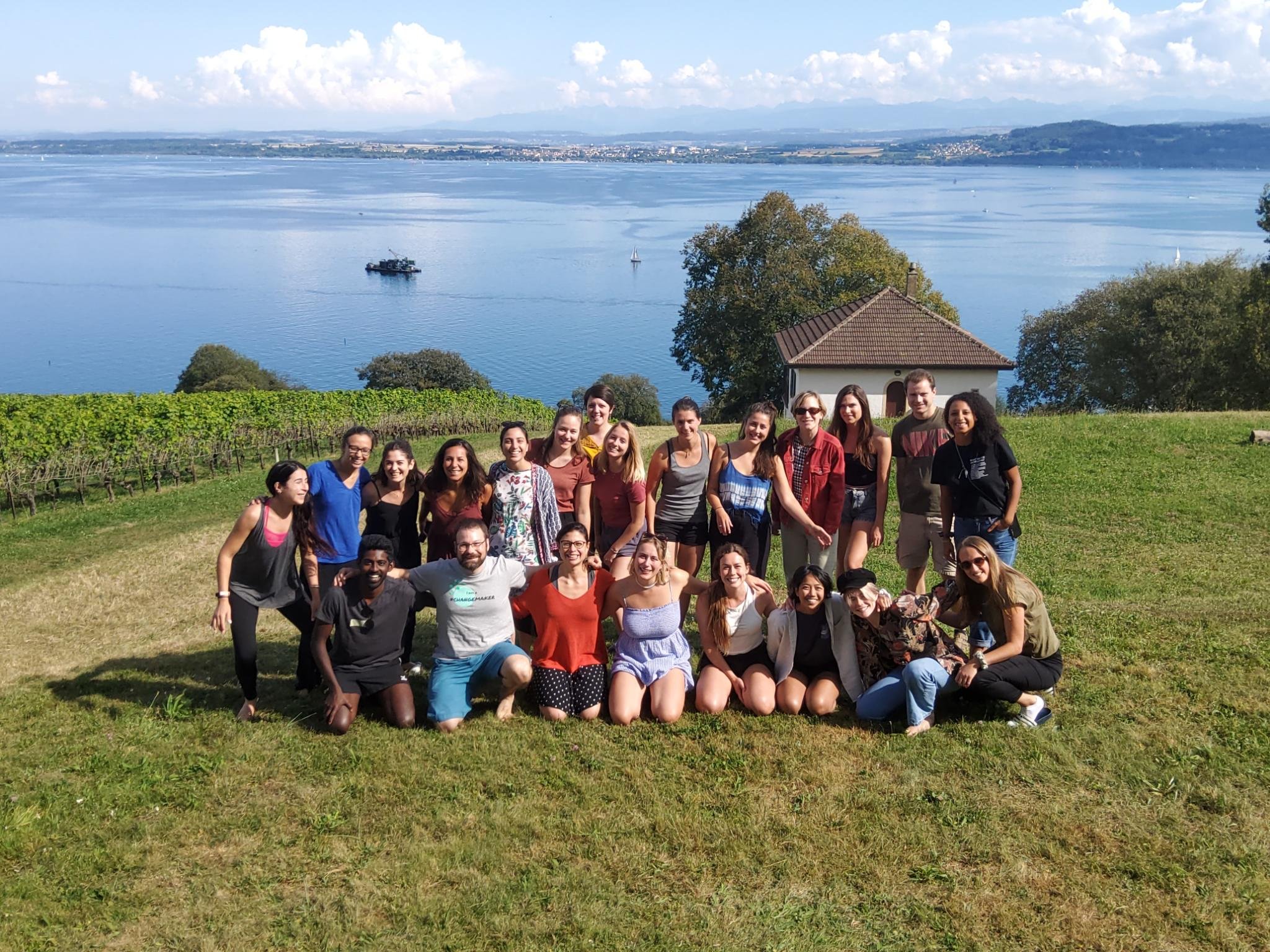 Gruppe von Menschen auf einer Wiese mit Blick auf einen See, im Hintergrund Weinreben und Berge.