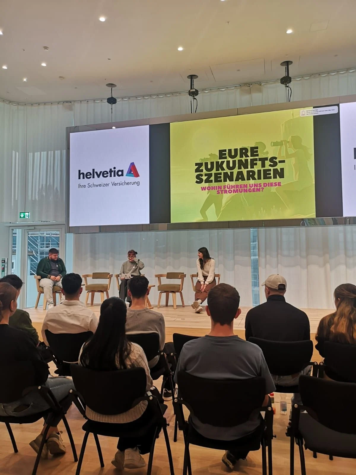 Podiumsdiskussion in einem Veranstaltungsraum mit einem großen Bildschirm im Hintergrund, der "Eure Zukunftsszenarien" zeigt. Mehrere Personen sitzen im Publikum, während drei Personen auf der Bühne diskutieren.