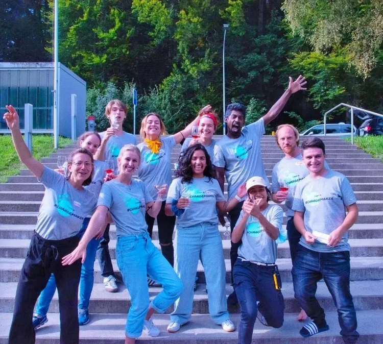 Gruppe von fröhlichen Menschen auf einer Treppe, die graue T-Shirts tragen, im Freien mit Bäumen im Hintergrund. Personen machen fröhliche Gesten und posieren zusammen. Es scheint ein sonniger Tag zu sein.