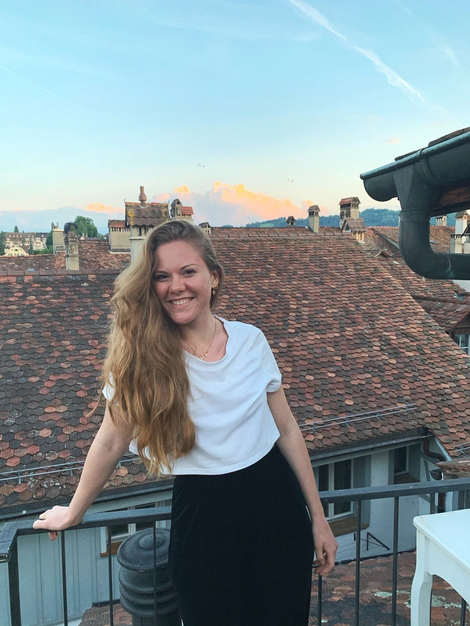 Frau auf einer Dachterrasse mit Ziegeldächern und blauem Himmel im Hintergrund.