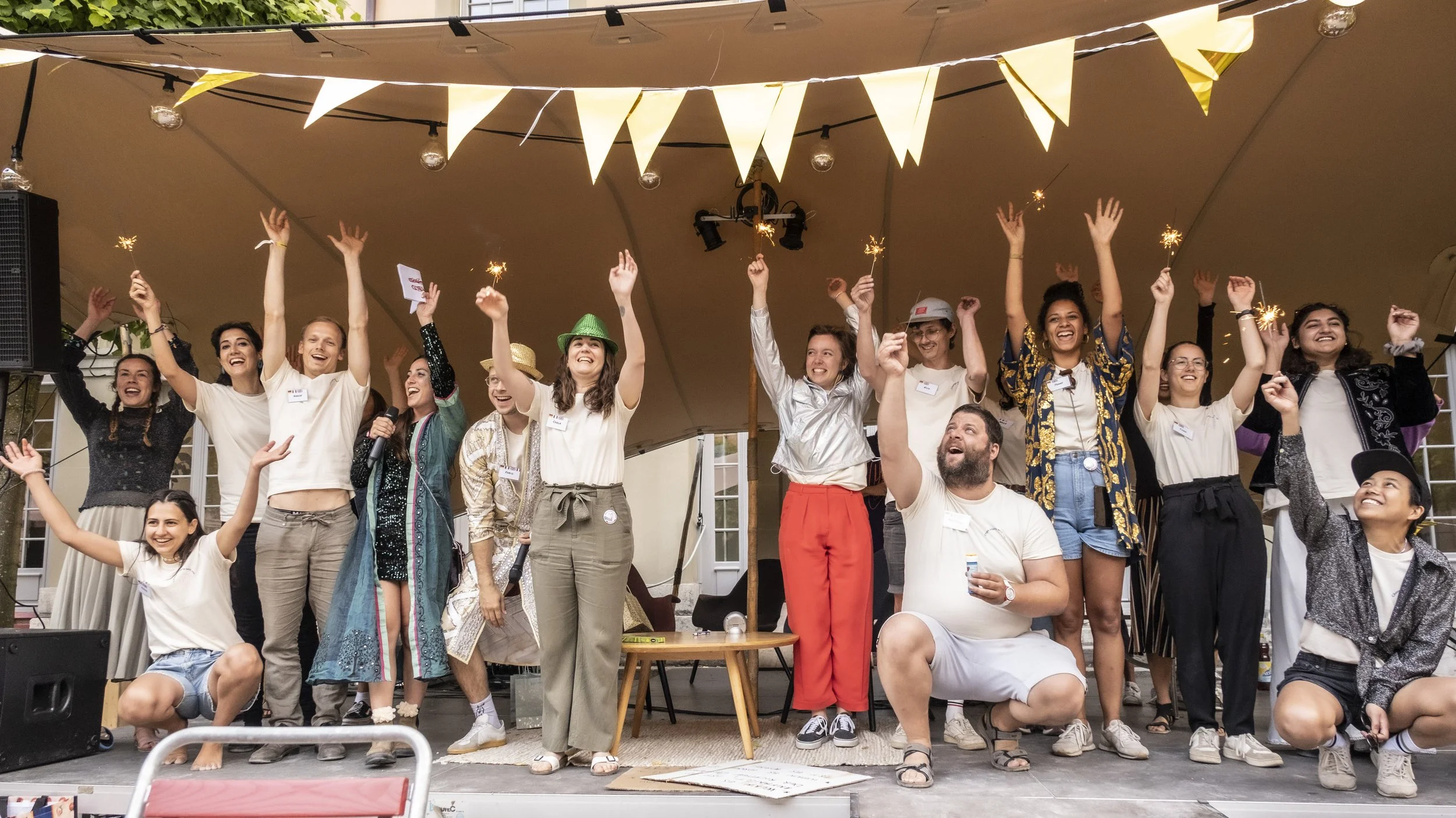 Eine Gruppe von Menschen bei einem Fest, fröhlich posierend auf einer Bühne. Sie heben die Arme und halten Wunderkerzen. Die Bühne ist mit gelben Wimpeln dekoriert.