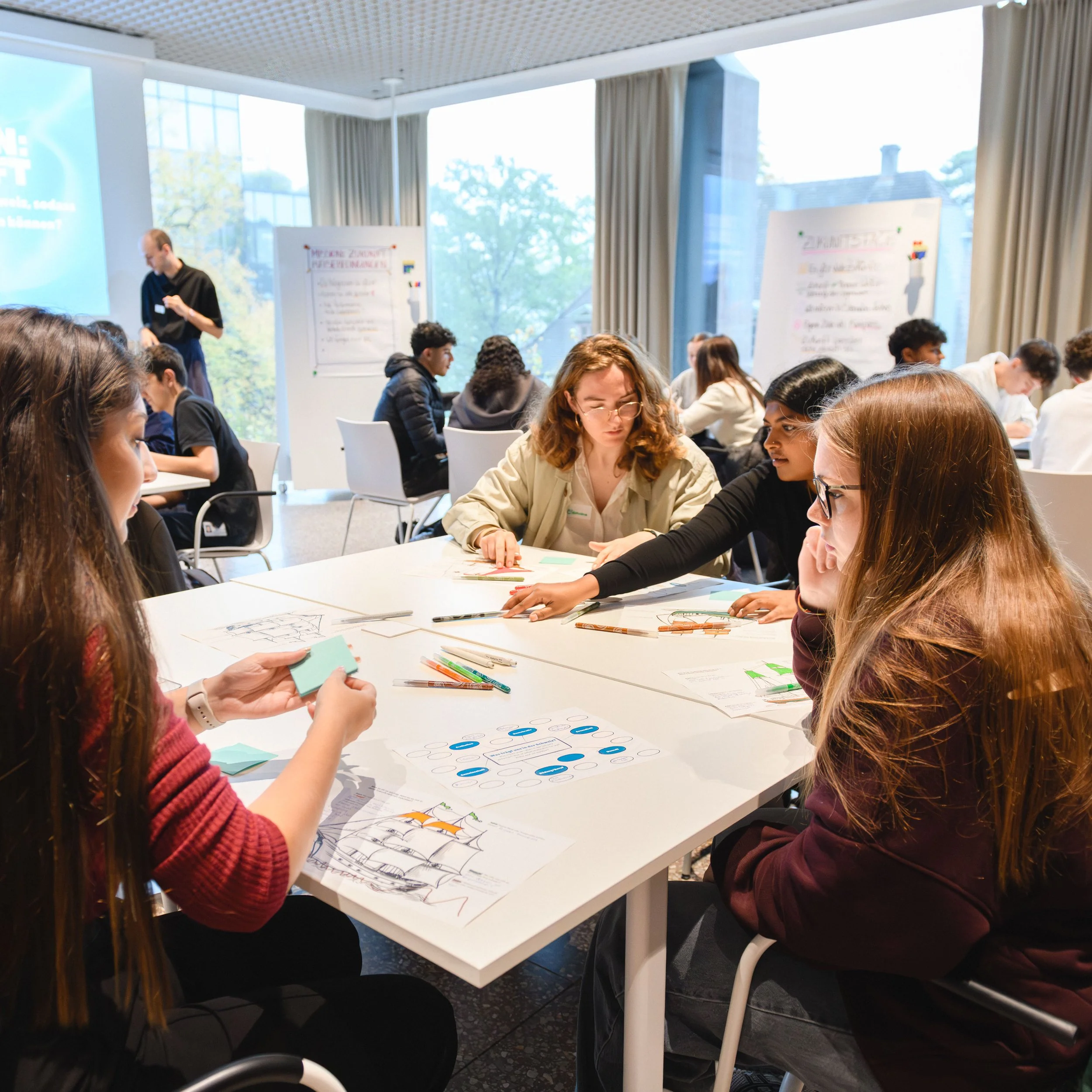 Podiumsdiskussion in einem Veranstaltungsraum mit einem großen Bildschirm im Hintergrund, der "Eure Zukunftsszenarien" zeigt. Mehrere Personen sitzen im Publikum, während drei Personen auf der Bühne diskutieren.