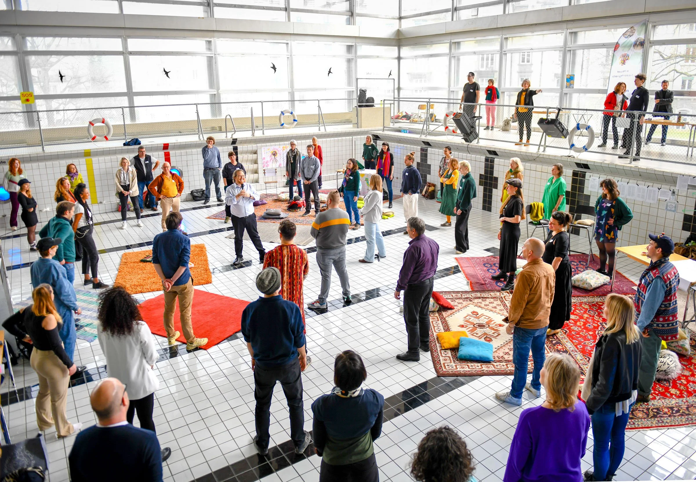 Eine Gruppe von Menschen steht im Kreis in einem leeren Schwimmbecken, das mit Teppichen bedeckt ist. Die Teilnehmer hören wahrscheinlich einer zentral stehenden Person zu, die spricht. Im Hintergrund sind weitere Personen und Schwimmring-Dekorationen zu sehen. Der Raum hat große Fenster.