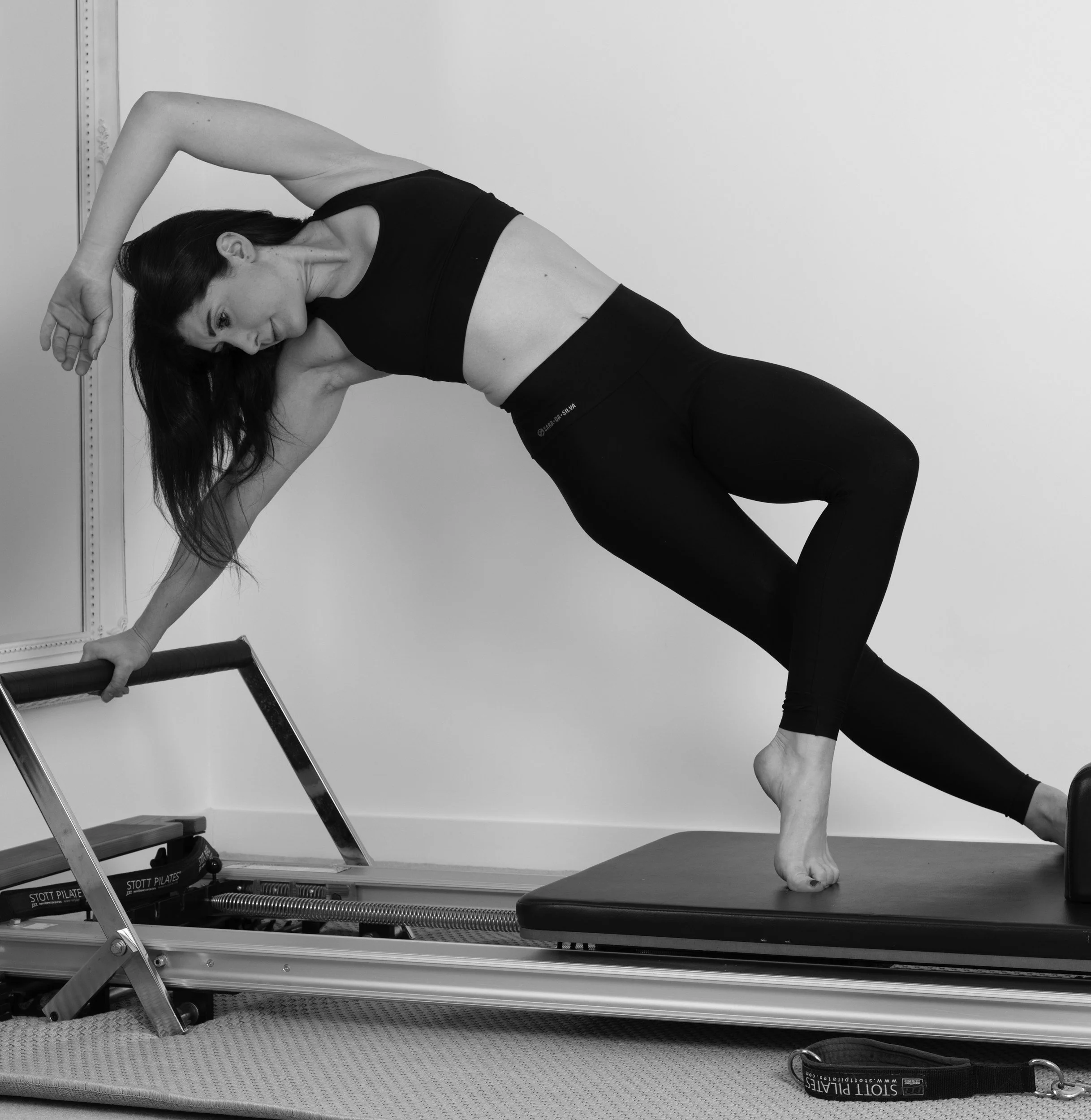 Een vrouw die yoga beoefent op een reformer pilates apparaat, met haar linkerhand op de handgreep en haar rechterbeen op een mat, in een pose waarbij ze haar linkerarm over haar hoofd buigt.
