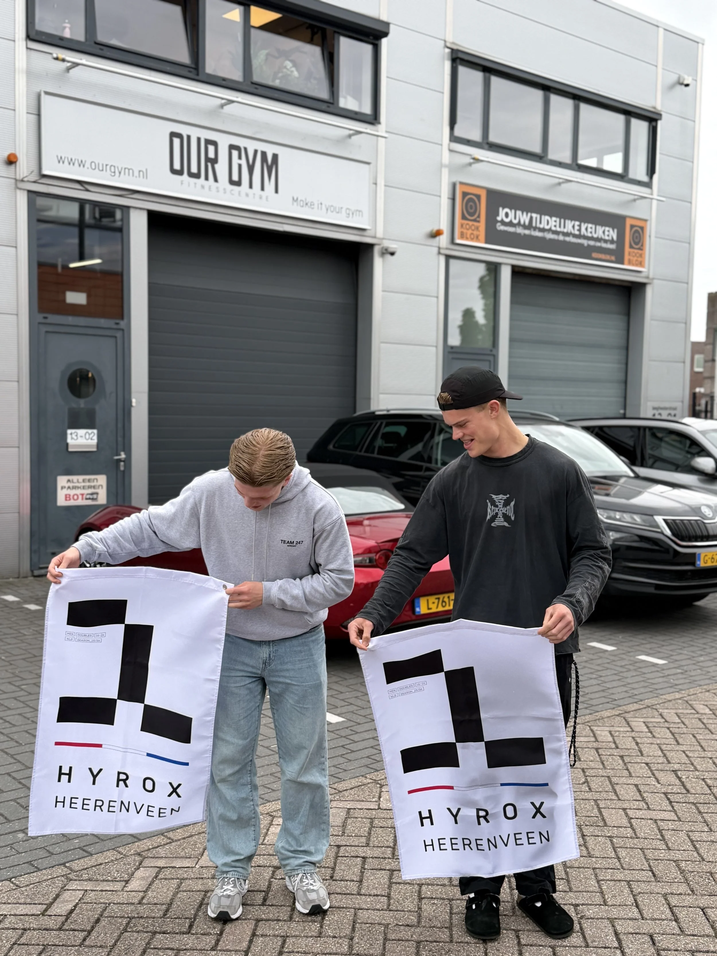 Twee jonge mannen houden banners vast met het logo van Hyrox Heerenveen, buiten voor een gebouw met ramen en een parkeergelegenheid. Ze lijken betrokken te zijn bij een evenement of actie.