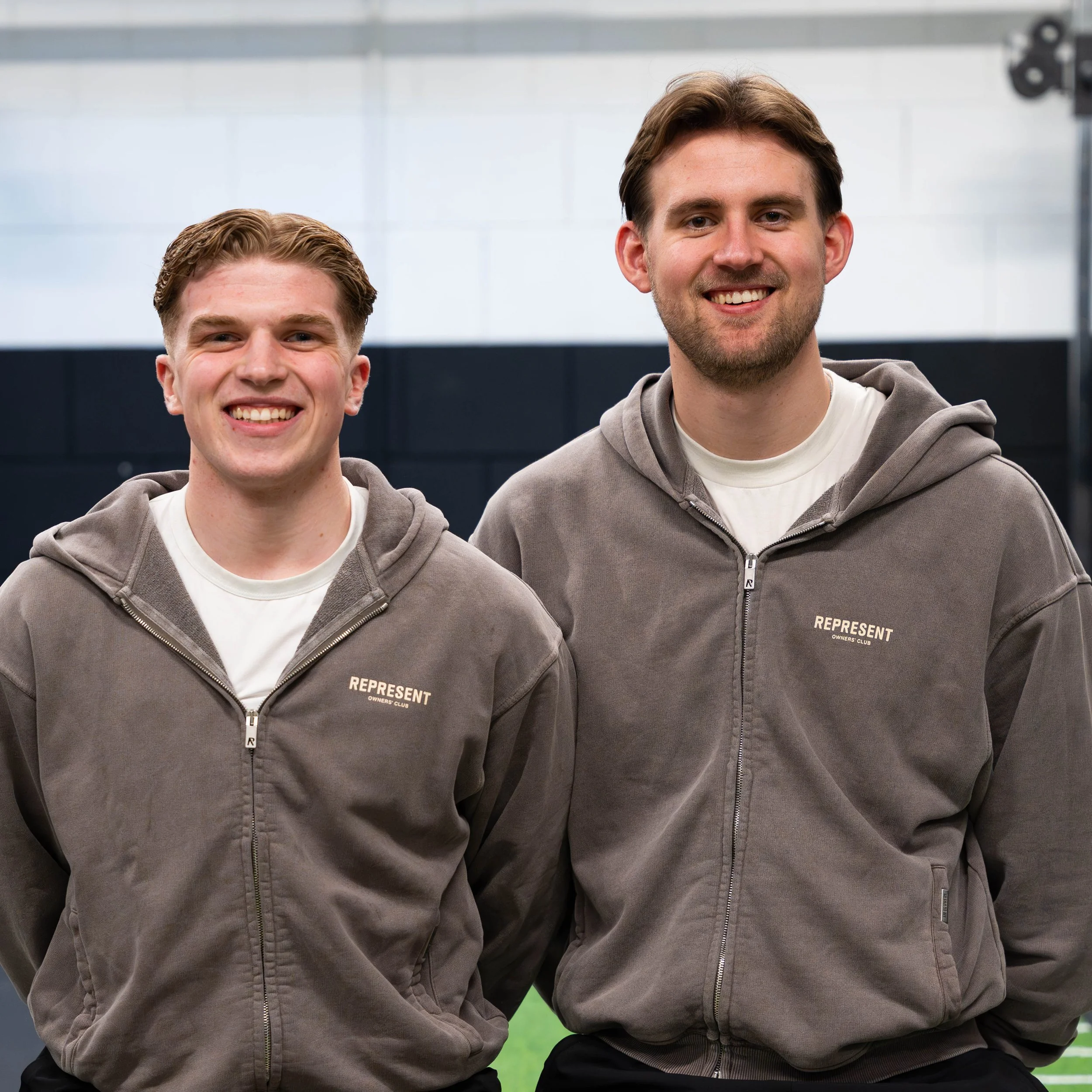 Een jonge man met een grijze hoodie die in een sportschool staat, lachend met de armen gekruist.
