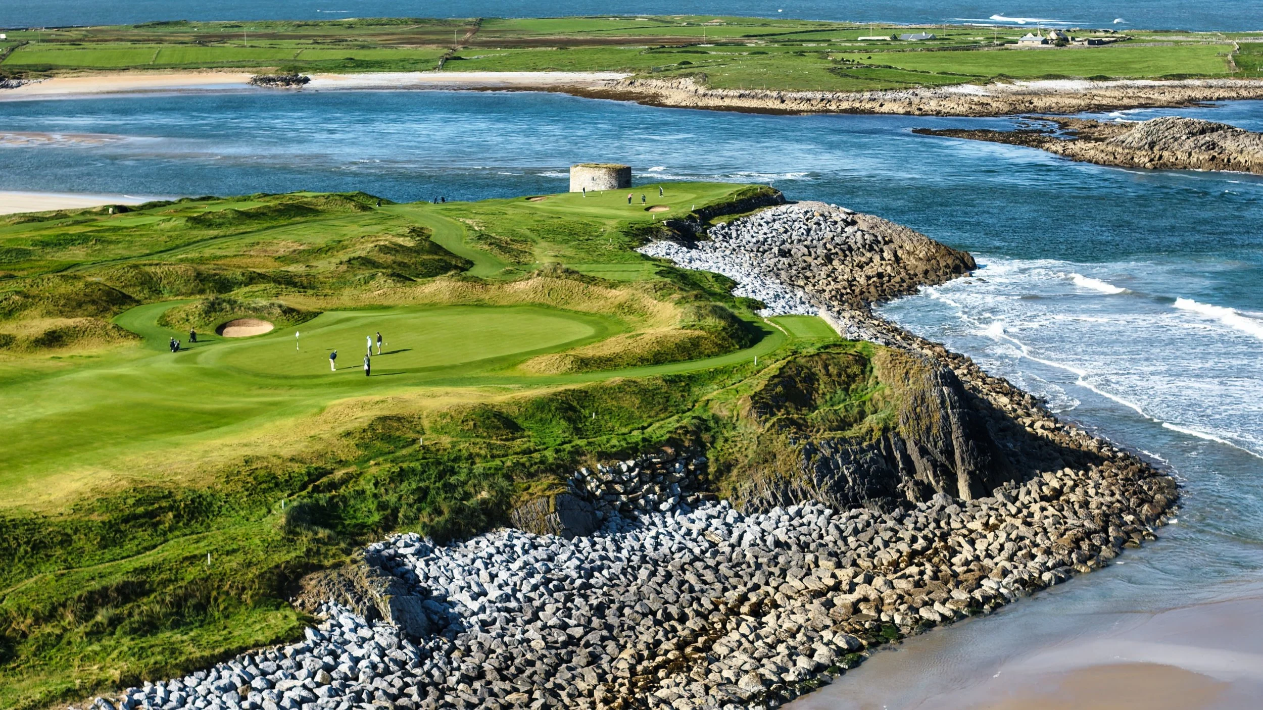 A scenic coastal golf course with players on the green, rugged rocks along the shoreline, and the ocean in the background.