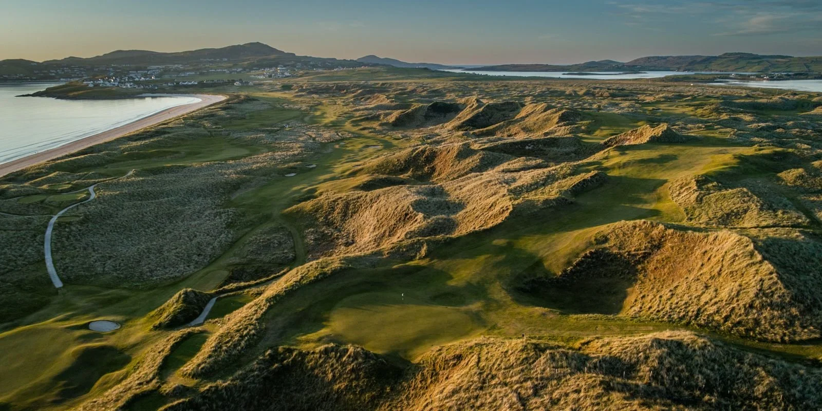 Aerial view of a golf course with green fairways, sand traps, and rolling hills beside a body of water, with mountains and a small town in the background.