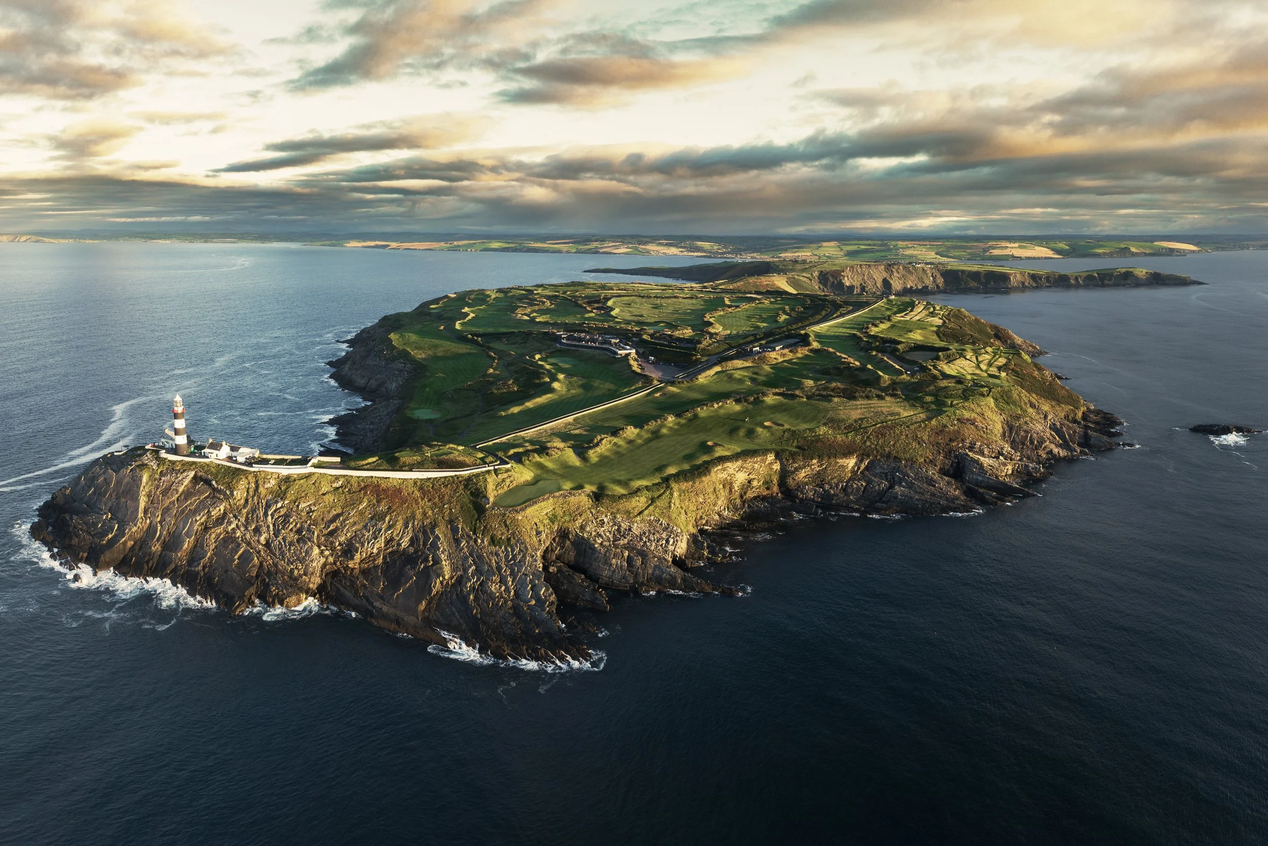 An aerial view of a lighthouse on a rocky coastline surrounded by green fields and tall cliffs, with the ocean and cloudy sky in the background.