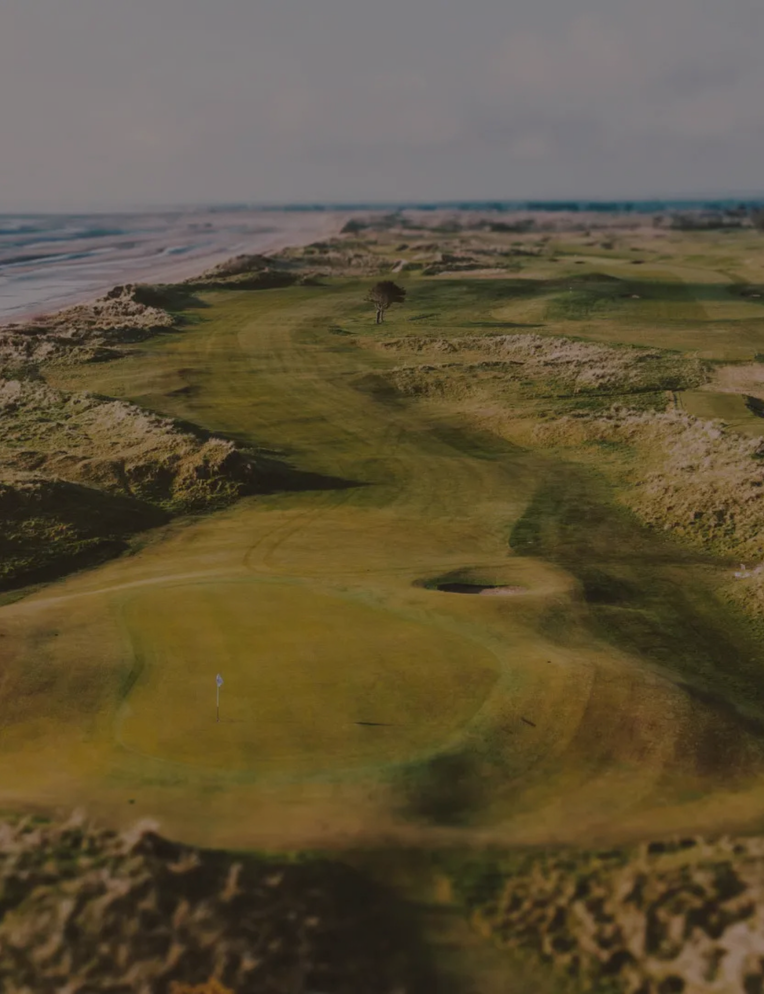 A golf course near a coastal area with a flag on the green, grass fairways, and ocean waves in the background.