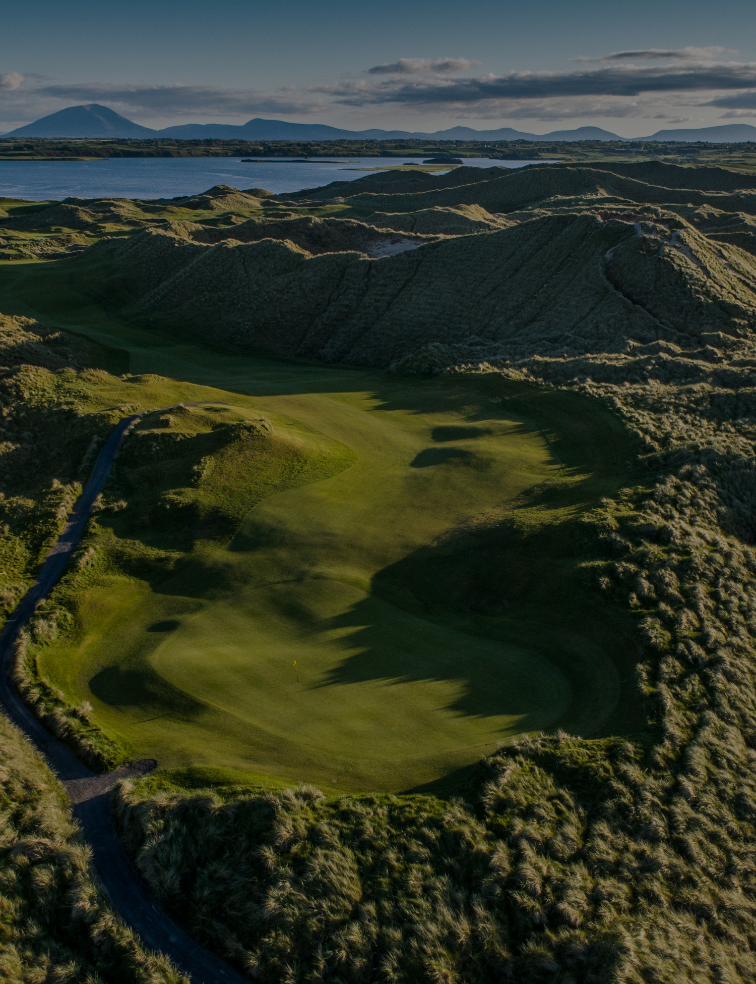 Aerial view of a golf course surrounded by grassy hills with a body of water and mountains in the background.