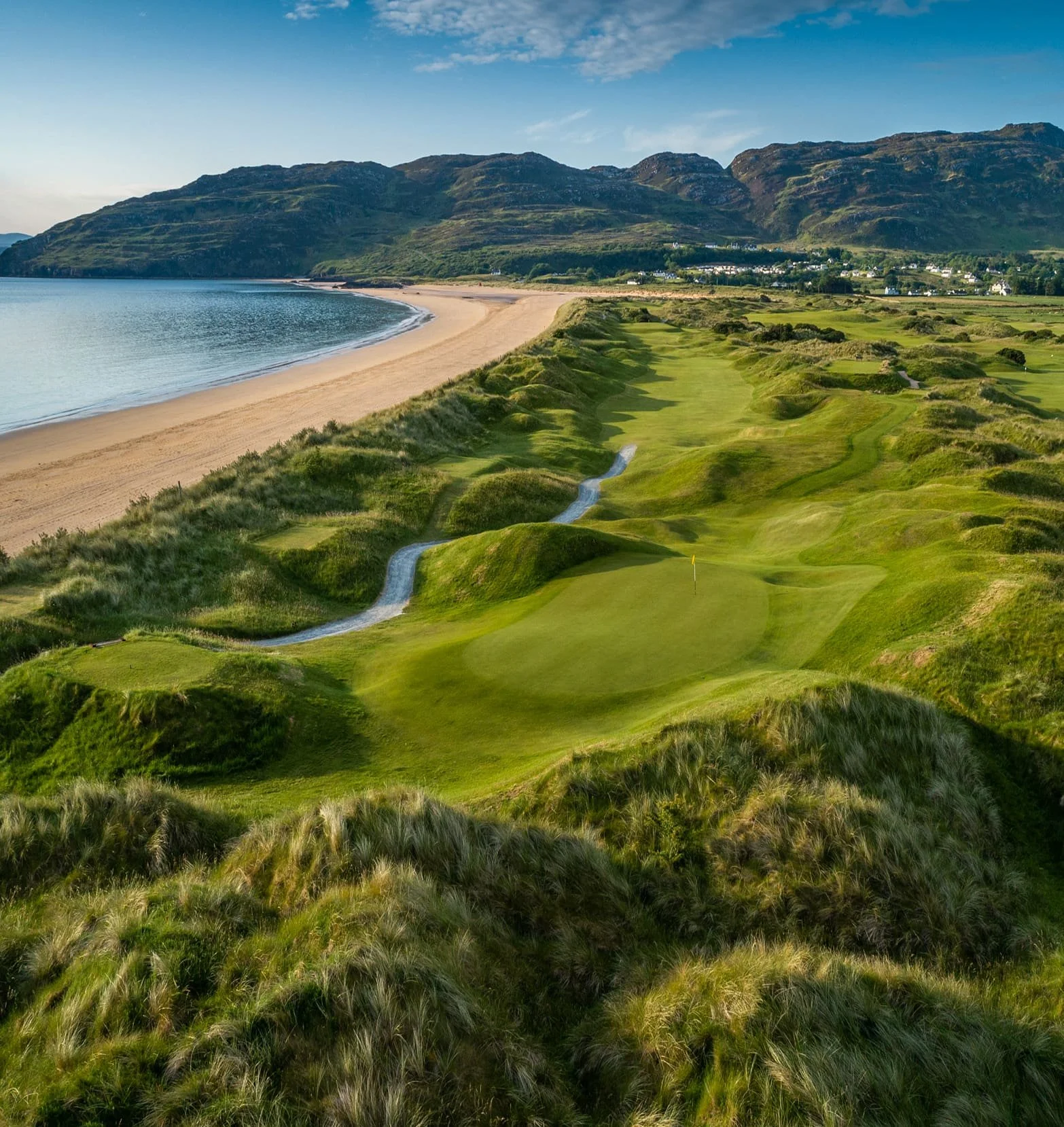 A scenic view of a golf course along a coastline with a sandy beach, hills, and mountains in the background under a blue sky with scattered clouds.
