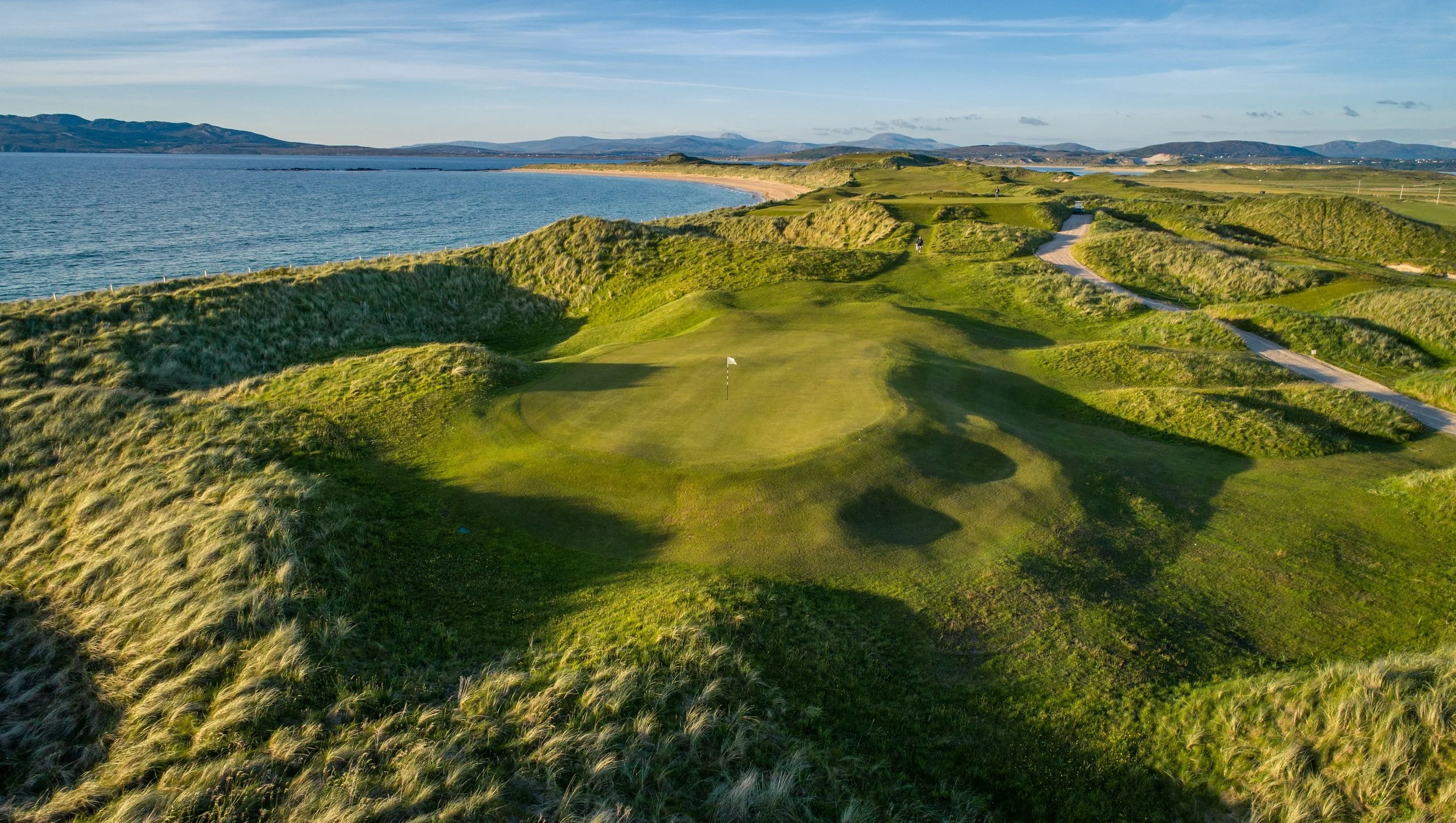 A scenic golf course located near a body of water with rolling green hills, sand traps, and a golf hole with a flag, under a clear sky.