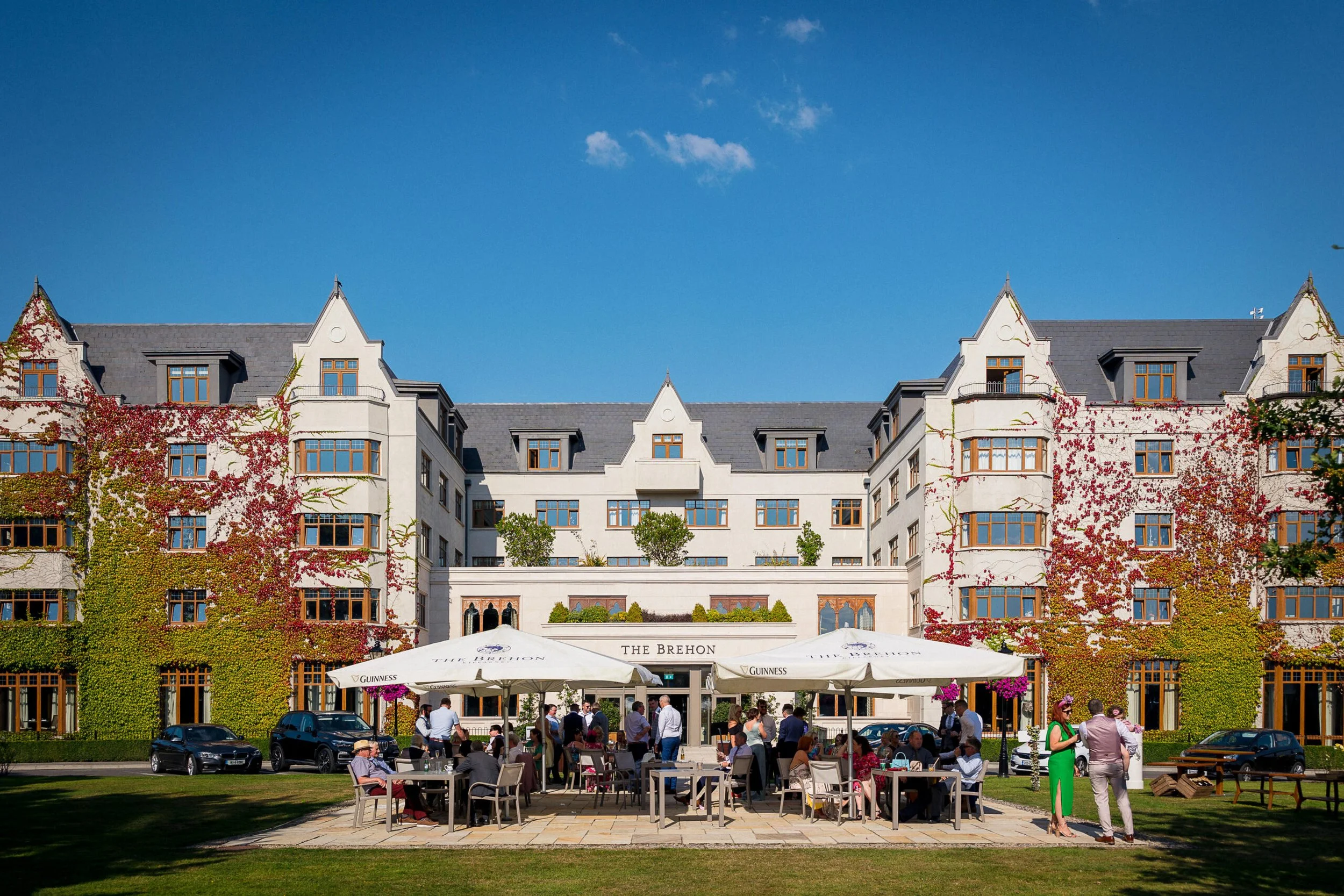 People dining outside at tables with umbrellas in front of a large white hotel building with ivy-covered walls, under a blue sky.