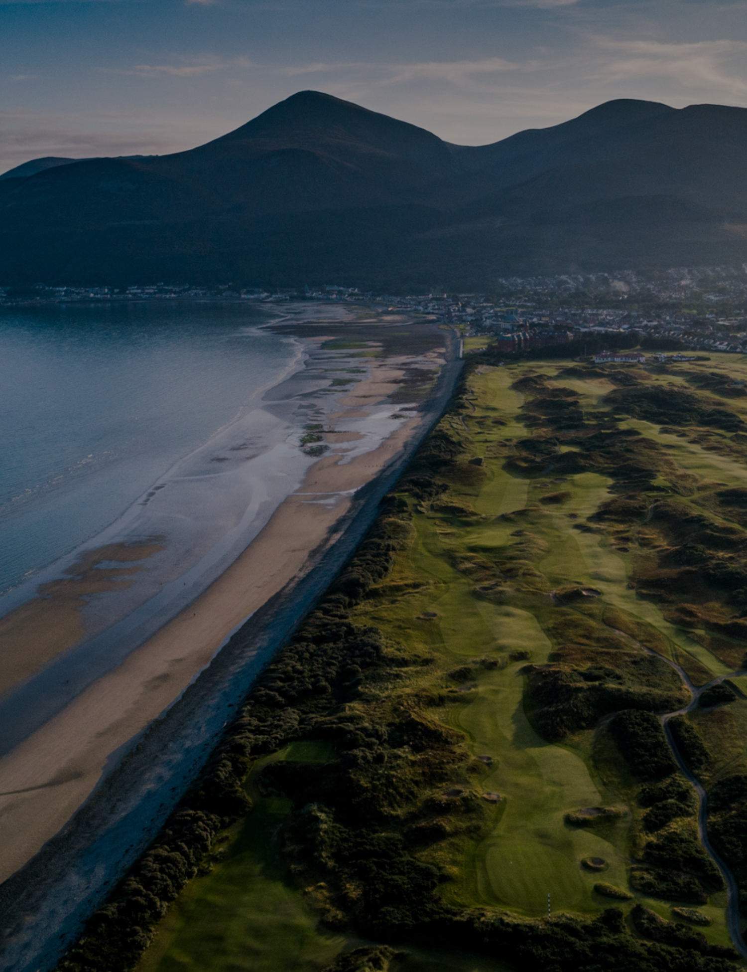Aerial view of a coastline with sandy beaches, green fields, and hills, with mountains in the background under a partly cloudy sky.