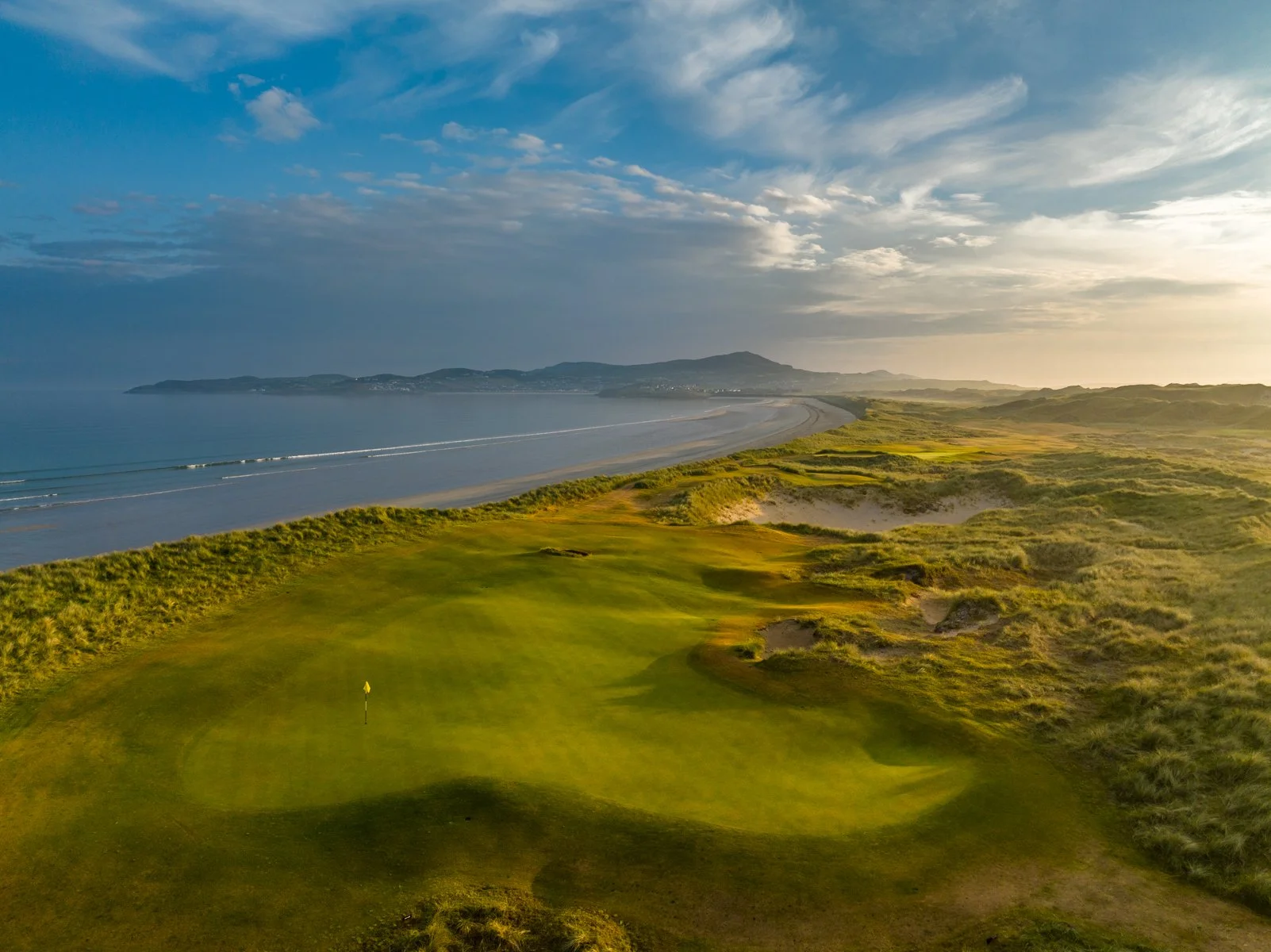 A golf course near the coastline with a green, a yellow flag, and sand dunes, overlooking the ocean and distant hills on a partly cloudy day.