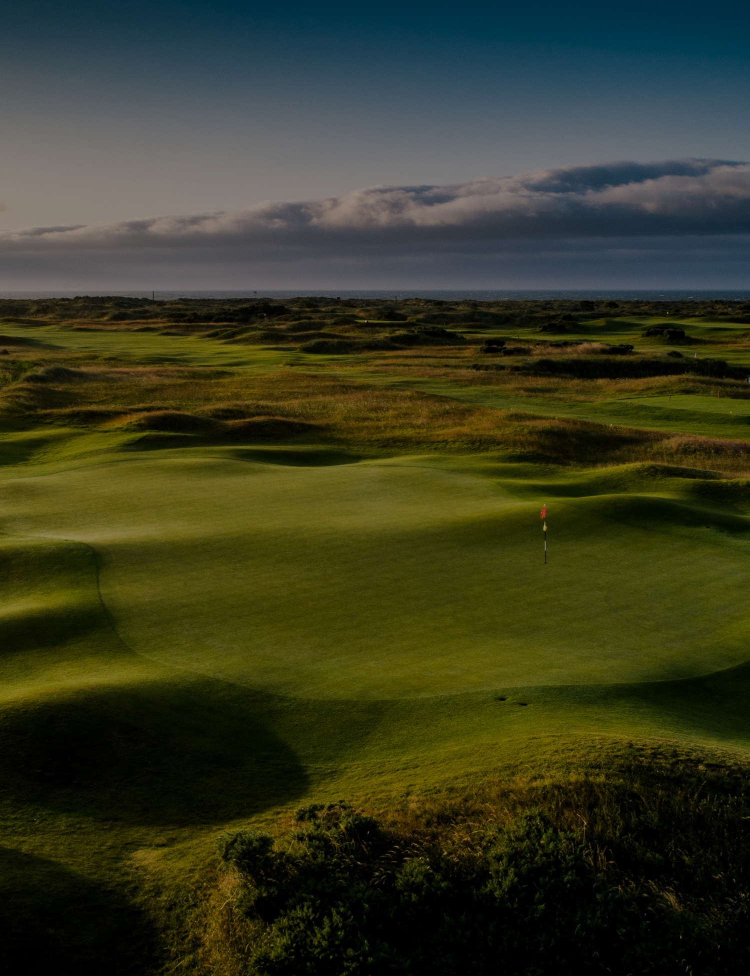 A landscape of a golf course at sunset with green fairways, sand traps, and a flagstick near the hole, under a partly cloudy sky.