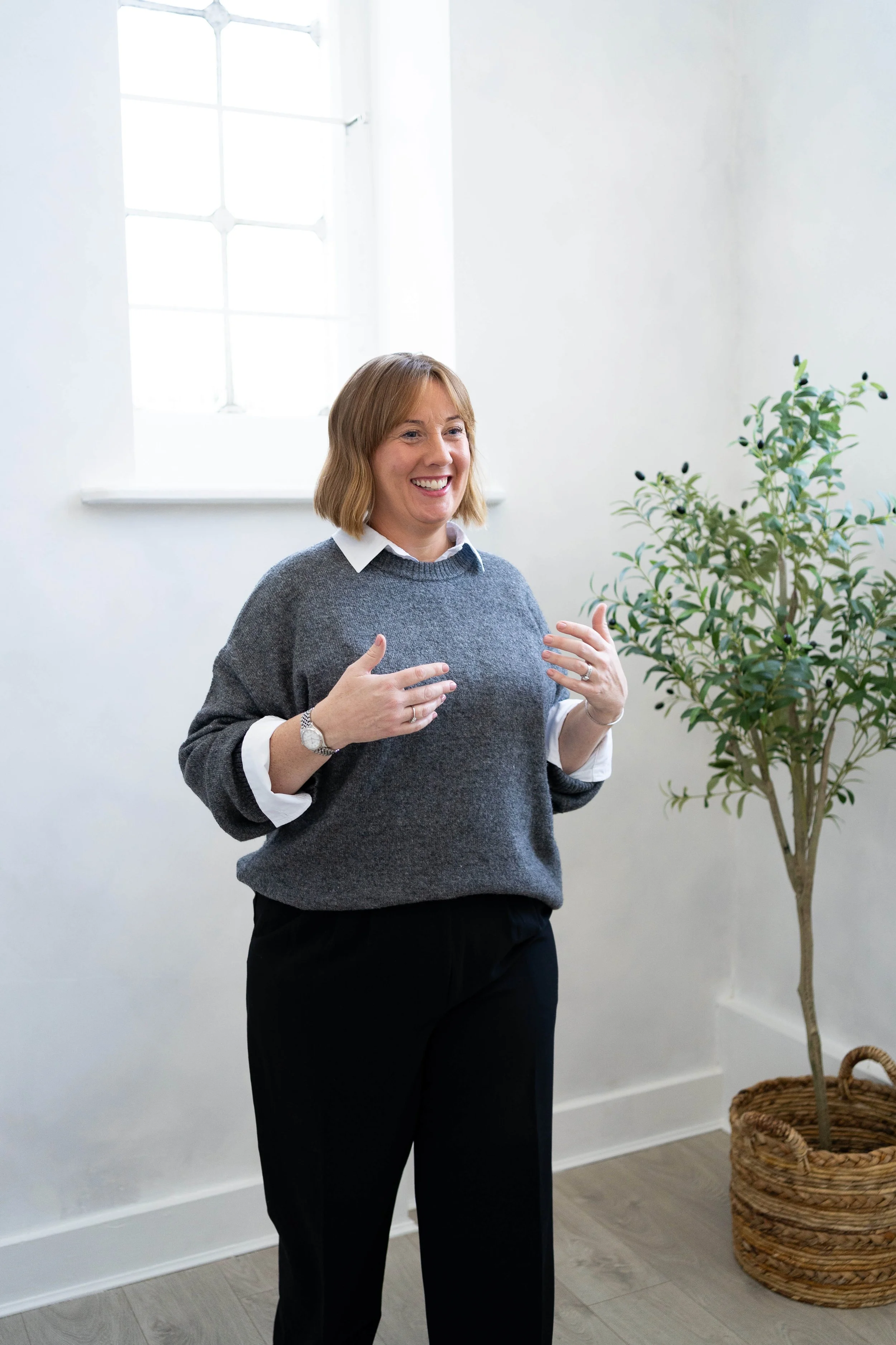 A woman with shoulder-length blonde hair, wearing a white shirt under a grey sweater and black pants, standing in a bright room with white walls, a window with decorative panes, and a potted plant in a woven basket. She is smiling and gesturing with her hands.