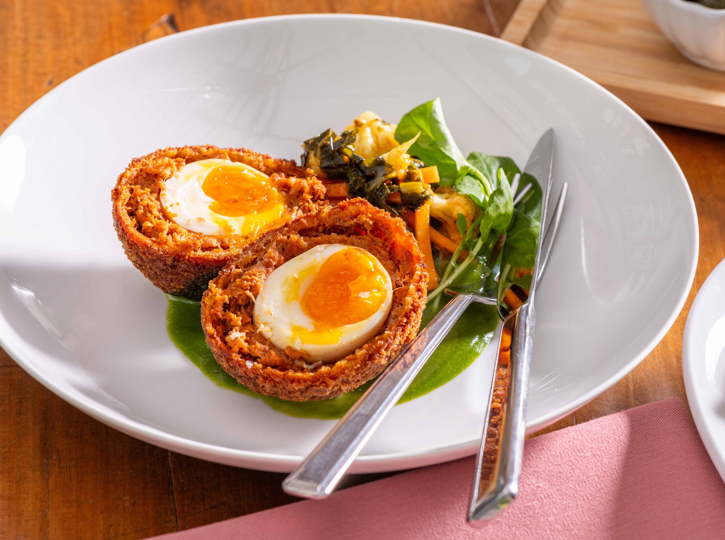 A white bowl containing two Scotch eggs with boiled eggs coated in seasoned meat and bread crumbs, sliced open to show runny yolks, garnished with a green sauce or puree. The bowl is on a wooden table with a fork resting on the edge and part of a pin