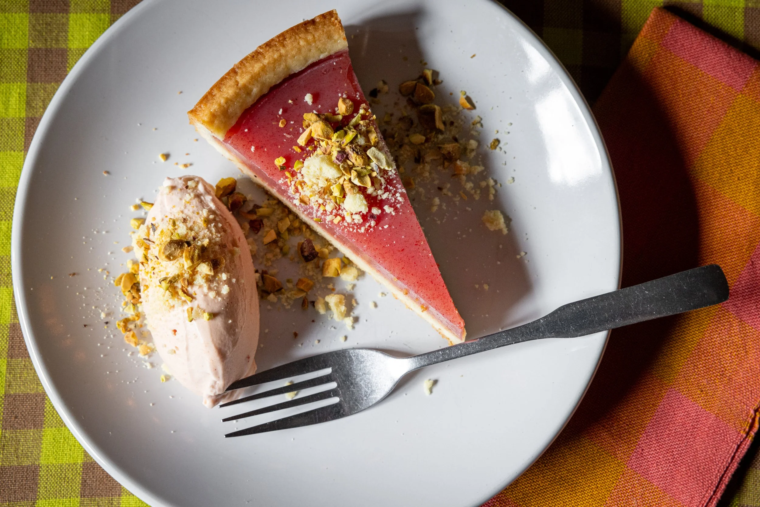 A slice of Sicilian watermelon tart topped with chopped pistachios, served on a white plate with a fork, on a checkered tablecloth.