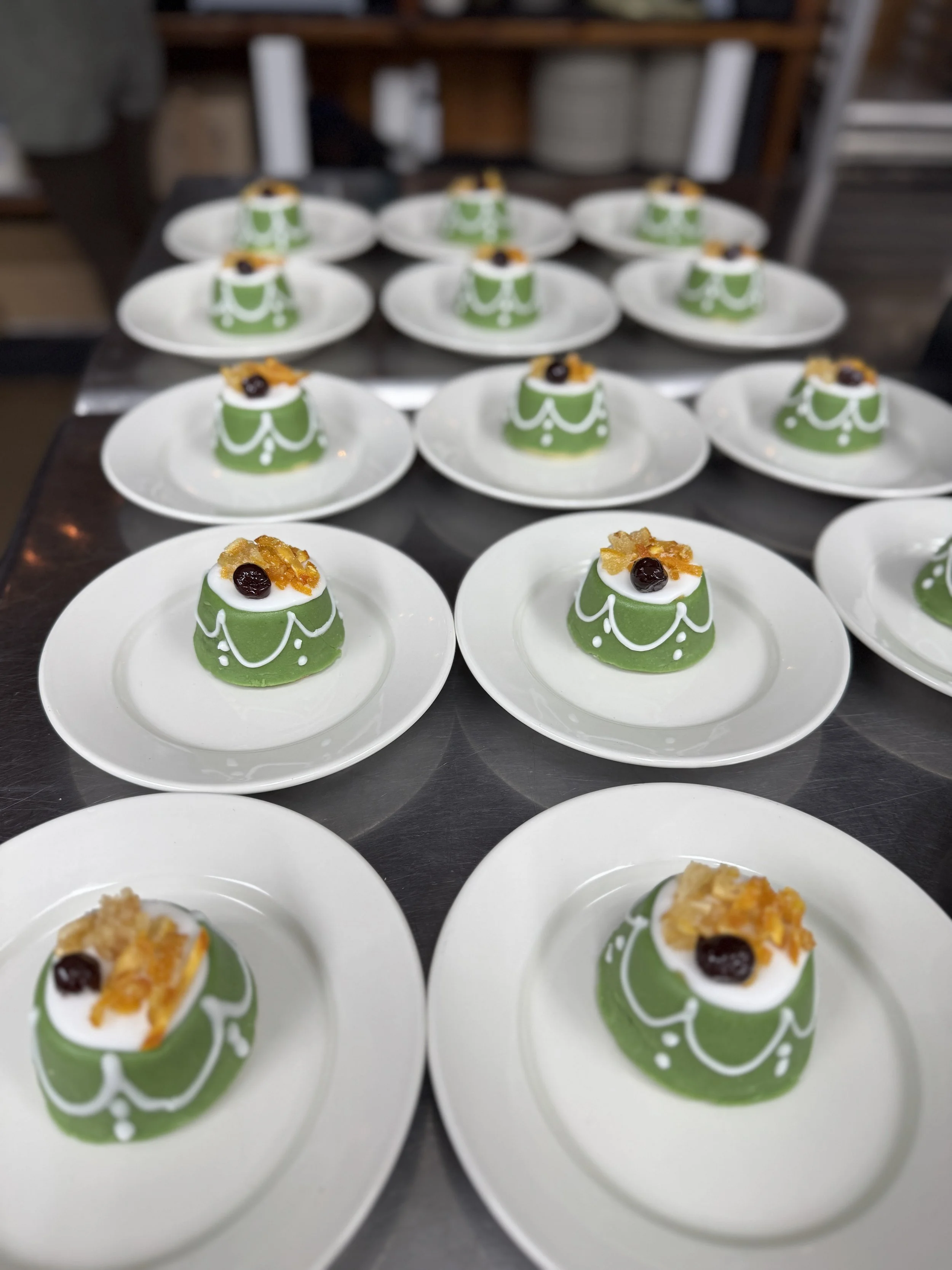 Multiple small green cakes decorated with white icing, topped with a cherry, orange peel, and a small dark fruit, arranged on white plates.