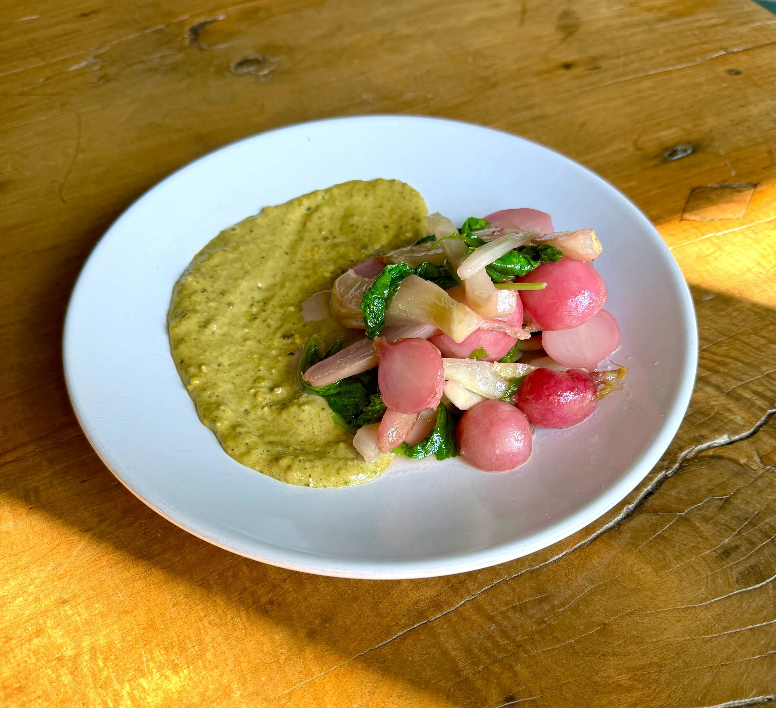 Close-up of a white plate containing pistachio butter, sliced and roasted radishes, and chopped green herbs on a wooden table.