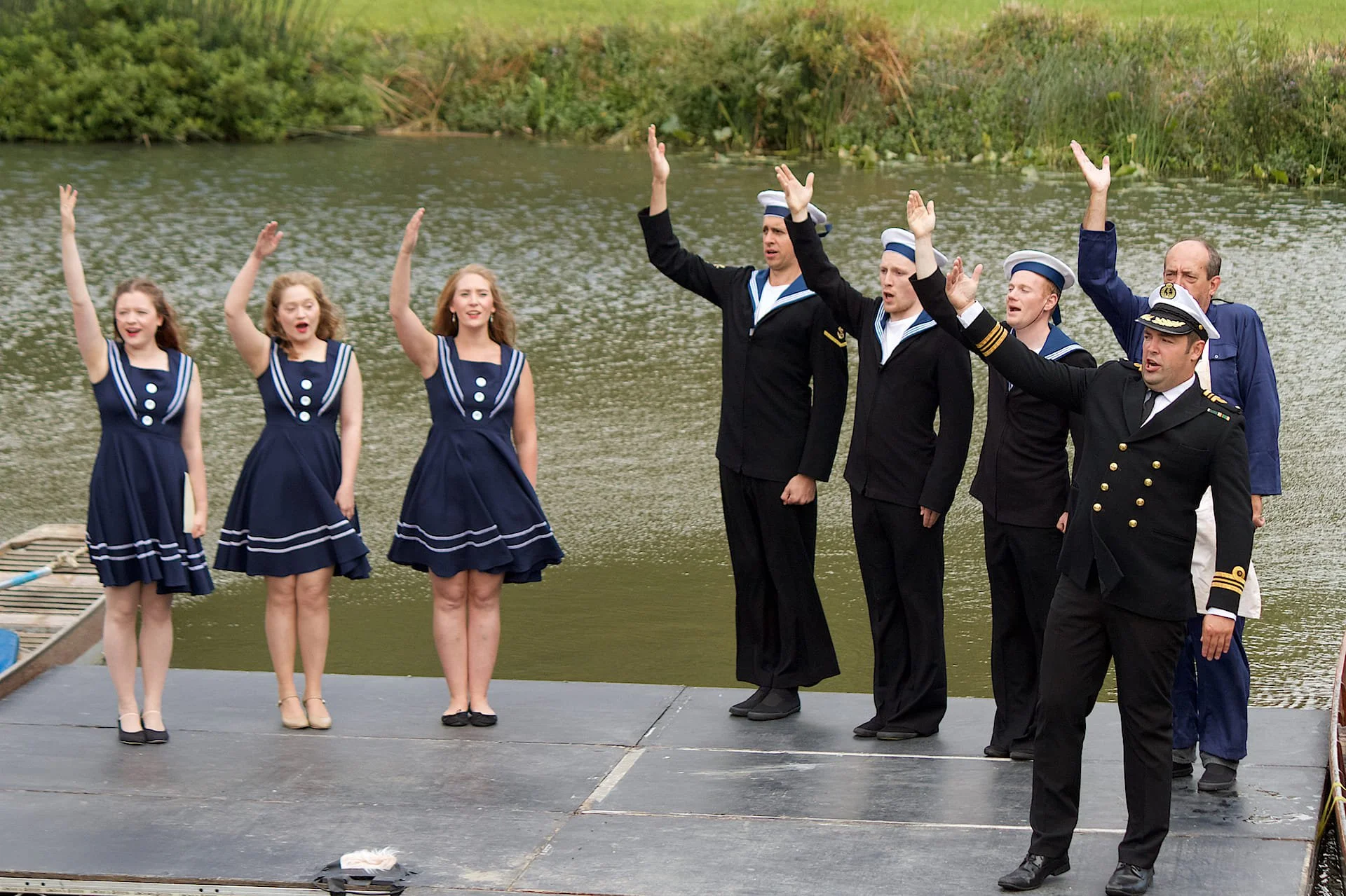 Women dressed in Navy sailor inspired dresses and men dressed as Navy sailors stand on a platform in front of a lake/pond with their right arms raised as they sing - opera anywhere