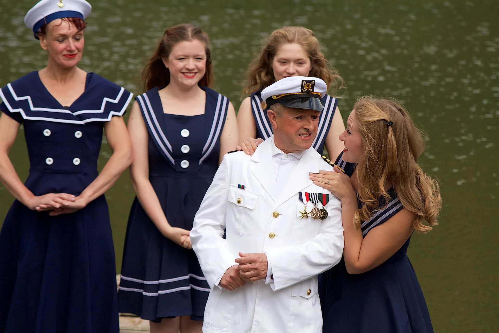 Man in a white Navy Officer uniform is spoken to by a woman in a navy-inspired dress. Three more women stand behind them. - opera anywhere