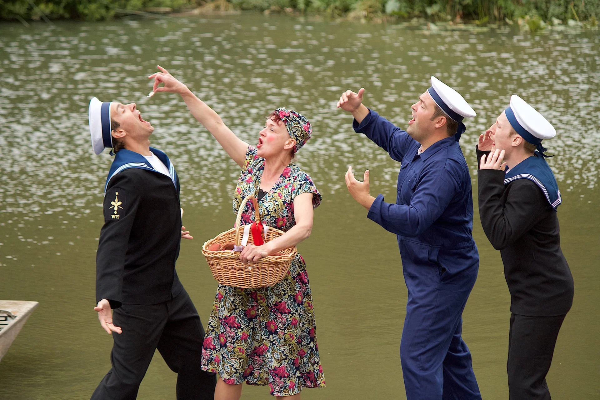 Woman in a flowery dress holding a woman basket feeds a man dressed as a Navy sailor who has his mouth wide open. Two other men stand by them, mid singing. - Opera Anywhere