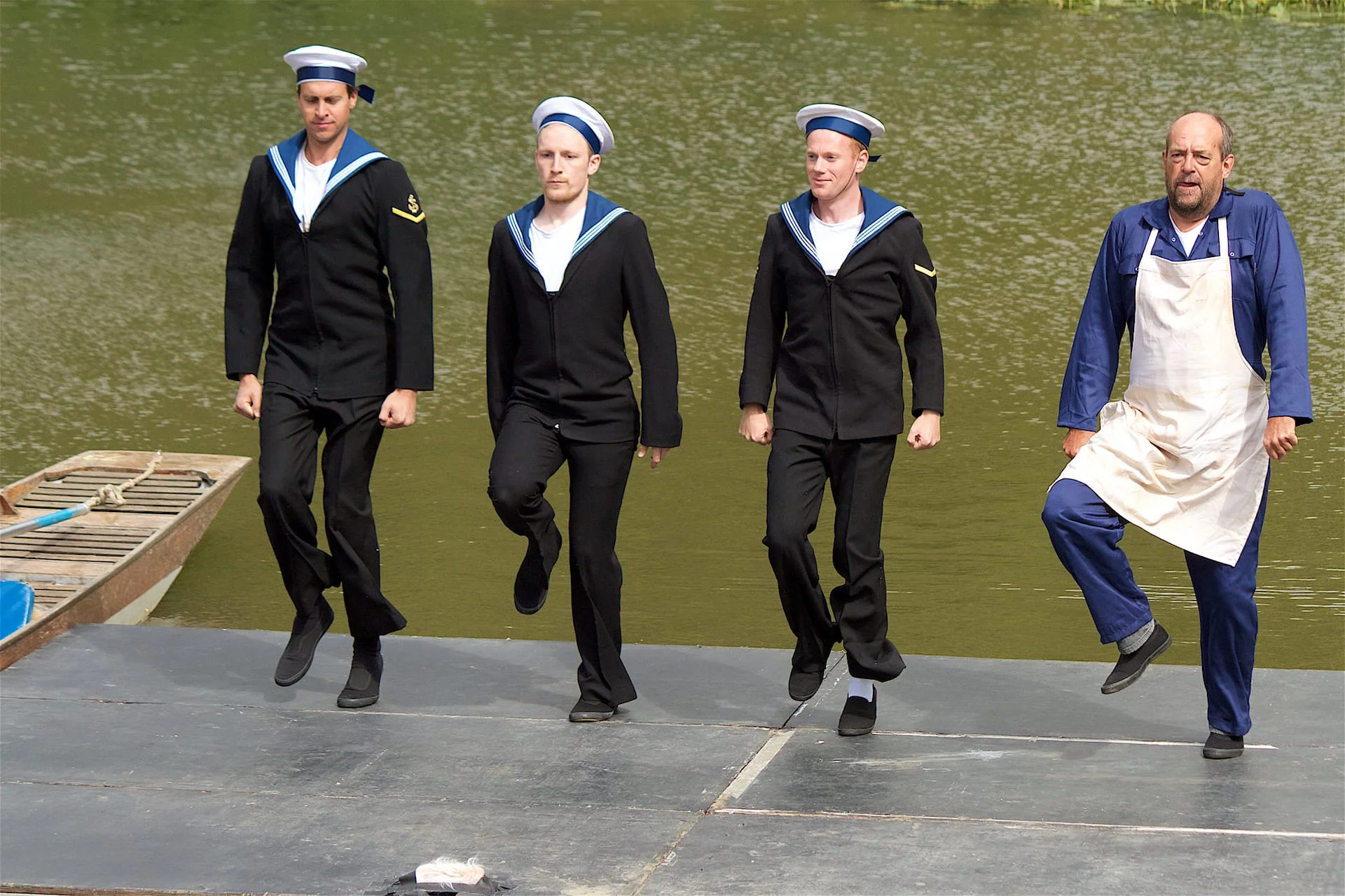 Three men dressed as Navy sailors (1940s style) and a man dressed as a navy cook dance on a platform in front of some water. They are mid air in a hop. - Opera Anywhere