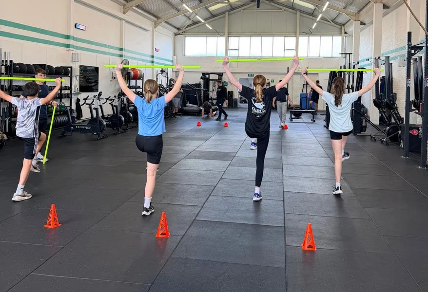 Female group fitness class balancing on one leg while holding green barbells in a gym